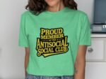 Woman wearing a green t-shirt with "PROUD MEMBER OF THE ANTISOCIAL SOCIAL CLUB" printed in bold black and yellow text.