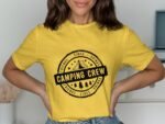 A woman wearing a yellow t-shirt with "Camping Crew" and outdoor-themed graphics, posing indoors.