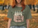 A woman wearing a green t-shirt featuring a graphic of bottles and the text "Just Here for the Boos." Autumn leaves in background.