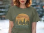 A woman wearing a green t-shirt with a sunset and pine trees graphic, against a blurred outdoor background.