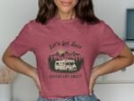 Woman wearing a pink shirt with "Let's Get Lost" graphic featuring a camper and mountains, smiling against a light background.