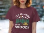 A person wearing a maroon t-shirt with “It's all good in the woods” graphic, featuring trees, mountains, and a tent.
