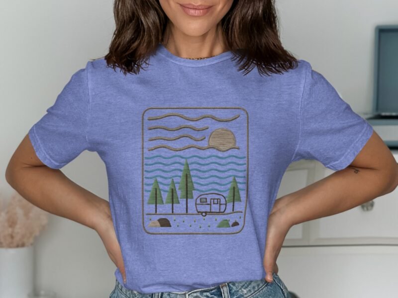 Woman wearing a light blue t-shirt with a vintage camping graphic of trees, water, a camper, and a sun.
