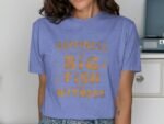 A woman wearing a lavender t-shirt with gold lettering that says "Happiness is a Big Fish and a Witness."