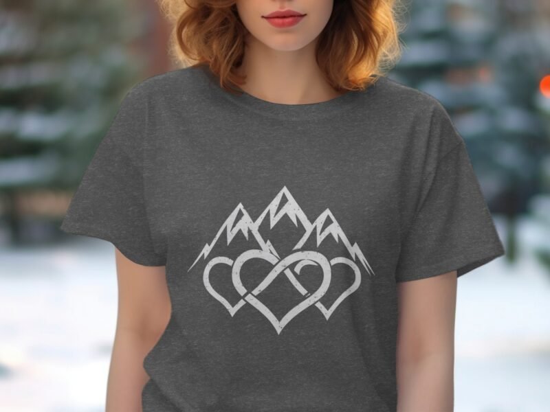 A woman in a gray t-shirt with a mountain and heart design stands outdoors in a wintry setting.