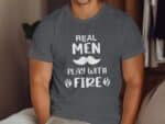A man in a gray t-shirt with the text "REAL MEN PLAY WITH FIRE" and a mustache graphic, sitting indoors.