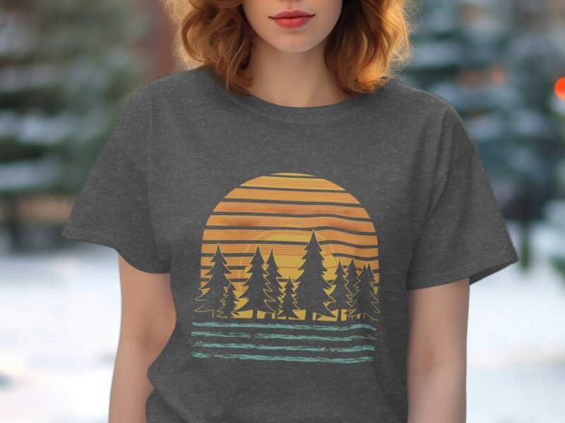 A woman in a gray t-shirt featuring a sunset with stripes, pine trees, and water, set against a blurred background.