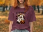 A woman with red hair wears a maroon T-shirt featuring a cartoon ghost cow with a pumpkin in a fall setting.