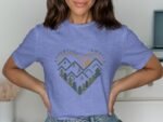 Woman in a light blue t-shirt with a heart design featuring mountains, trees, and the text "Adventure Awaits."