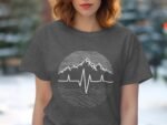A woman with wavy hair wears a dark gray t-shirt featuring a mountain and heartbeat design, with a blurred outdoor background.