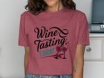 Woman wearing a maroon t-shirt with "Wine Tasting T-Shirt" graphic and wine glasses design. Background features furniture.