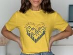 Model wearing a yellow t-shirt with a heart-shaped mountain graphic and the text "Adventure Awaits." Background is neutral.