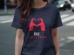 A woman in a navy t-shirt featuring a red design of two birds and the text "BE YOUTIFUL" stands on a city street.