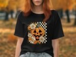 A person wearing a black t-shirt featuring a pumpkin design with daisies and a checkered background, set outdoors.