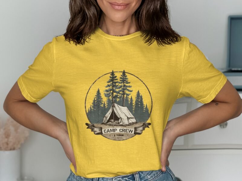 A woman models a yellow t-shirt featuring a camping design with trees and a tent, indoors against a neutral background.
