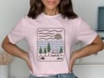 Woman wearing a pink t-shirt with a graphic of trees, a camper, waves, and the sun illustrated.