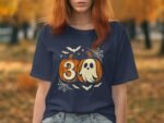 A woman in a navy t-shirt featuring a design with pumpkins, a ghost, and the number 30, surrounded by bats and spider webs.