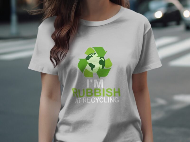 A woman wearing a gray t-shirt with a recycling logo and the text "I'M RUBBISH AT RECYCLING" stands on a crosswalk.