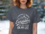 A woman in a gray t-shirt with the text, "I Haven't Been Everywhere But It's On My List," against a blurred outdoor background.