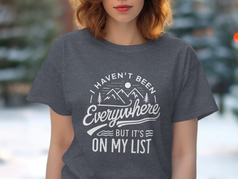 A woman in a gray t-shirt with the text, "I Haven't Been Everywhere But It's On My List," against a blurred outdoor background.