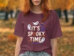 A woman with reddish hair wears a maroon t-shirt featuring a Halloween design that says "It's Spooky Time."