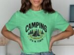 A woman wearing a mint green t-shirt with a camping design featuring a tent and trees, posing with hands on her hips.