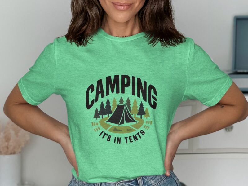 A woman wearing a mint green t-shirt with a camping design featuring a tent and trees, posing with hands on her hips.