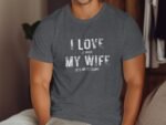 A man in a gray t-shirt with white text reading "I LOVE IT WHEN MY WIFE LETS ME GO FISHING" sits against a wooden background.