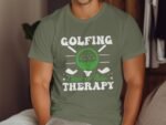 A person wearing an olive green t-shirt displaying the text "Golfing Cheaper Than Therapy" with golf graphics.