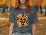 A woman in a gray t-shirt featuring a Halloween pumpkin design with daisies and a checkered background, set outdoors.