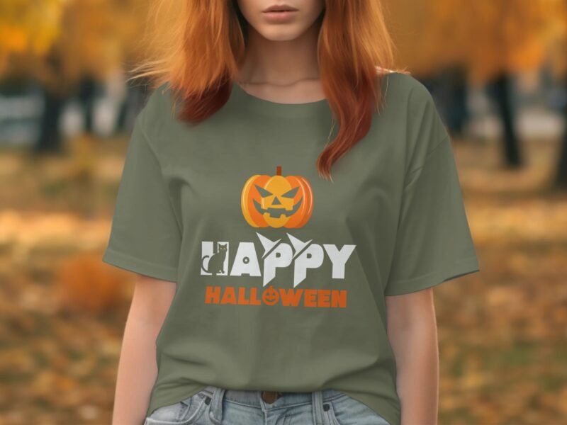A woman wearing a green t-shirt with “Happy Halloween” and a pumpkin graphic stands in a park with autumn foliage.