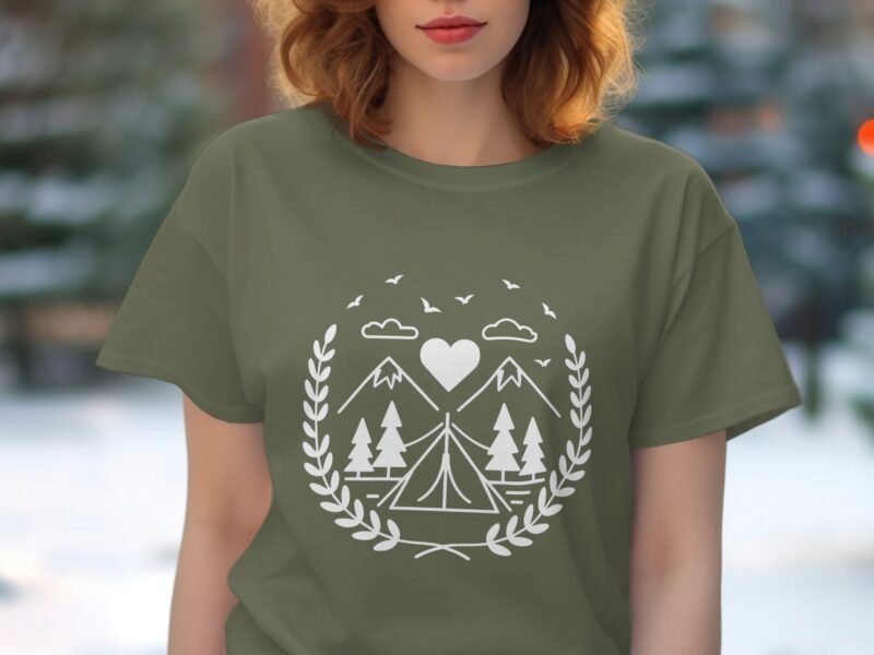 A woman wearing an olive green t-shirt with a white camping and mountain graphic design, outdoors.