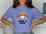 A woman wearing a lavender t-shirt with a sunset and "TAKE A HIKE" graphic, posing with hands on her hips.
