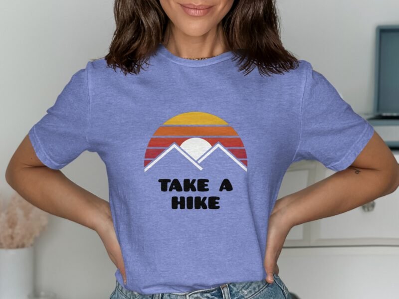 A woman wearing a lavender t-shirt with a sunset and "TAKE A HIKE" graphic, posing with hands on her hips.