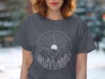 A woman wearing a dark gray t-shirt with a white mountain and sun graphic design, trees in foreground, outdoors setting.