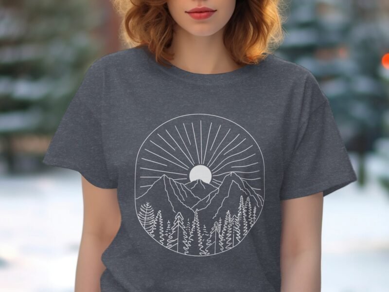 A woman wearing a dark gray t-shirt with a white mountain and sun graphic design, trees in foreground, outdoors setting.