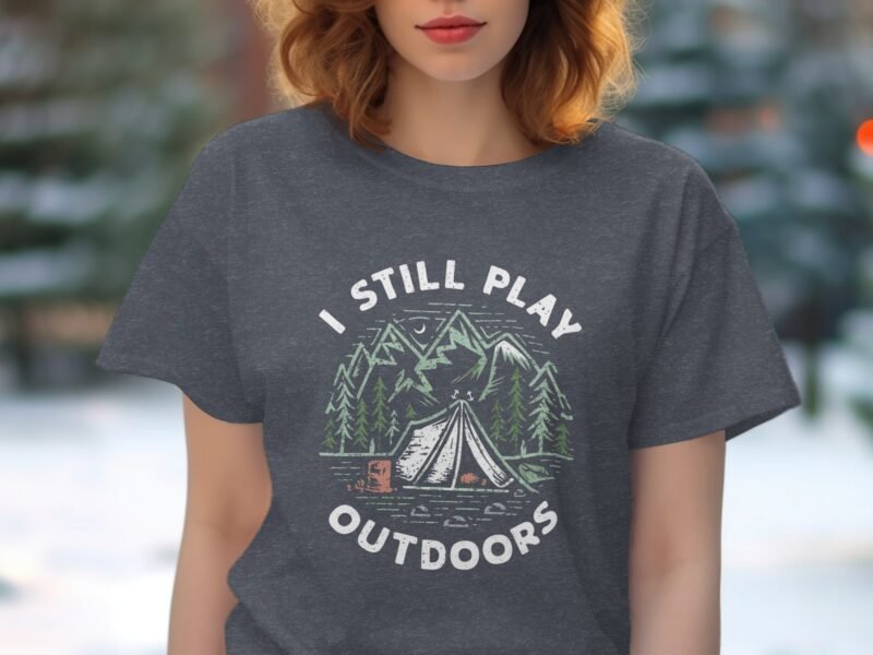 Woman wearing a gray t-shirt with "I STILL PLAY OUTDOORS" and a camping scene graphic, set against a blurred green background.