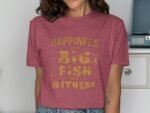 Woman wearing a maroon t-shirt with yellow text reading "Happiness is a Big Fish and a Witness" in a well-lit room.