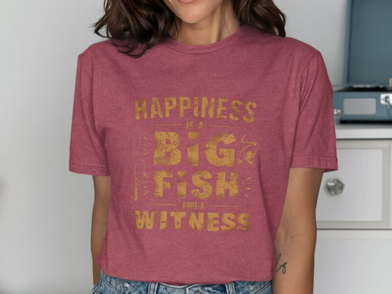 Woman wearing a maroon t-shirt with yellow text reading "Happiness is a Big Fish and a Witness" in a well-lit room.