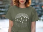 A woman in an olive green t-shirt featuring a mountain and heartbeat graphic, with a blurred outdoor background.