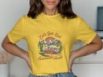 A woman in a bright yellow shirt featuring a vintage camper van design, surrounded by mountains and trees.