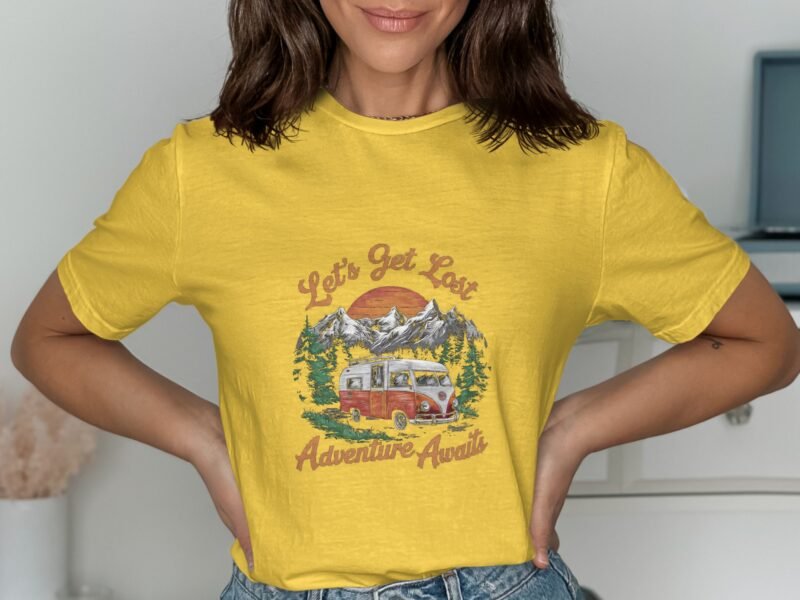 A woman in a bright yellow shirt featuring a vintage camper van design, surrounded by mountains and trees.