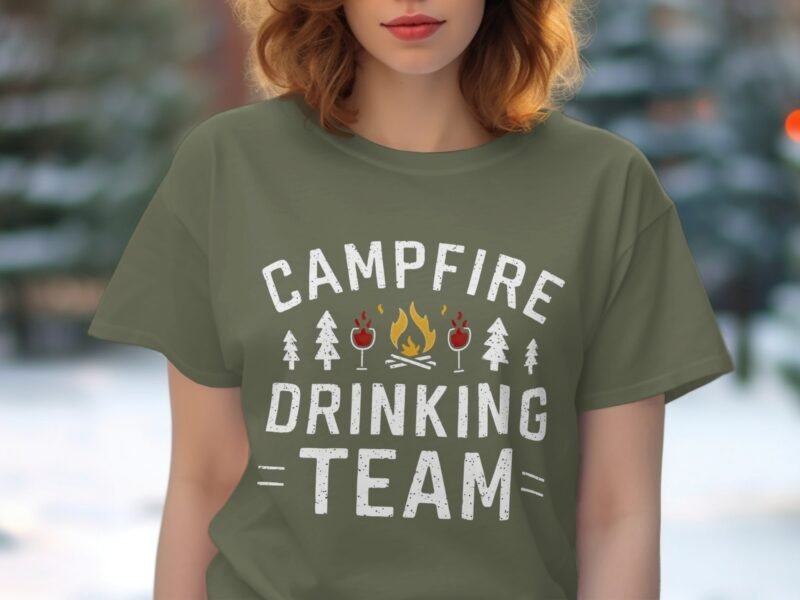 Woman wearing an olive green t-shirt that reads "Campfire Drinking Team," surrounded by trees in a blurred background.