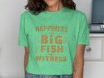 Woman wearing a green t-shirt with the phrase "HAPPINESS IS A BIG FISH AND A WITNESS" in orange text.