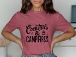 Woman wearing a pink t-shirt with the phrase “Cocktails & Campfires” in black font, smiling and posing.