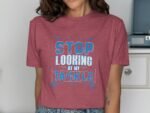 Woman wearing a maroon t-shirt with the text "STOP LOOKING AT MY TACKLE" in blue and white, with a casual background.