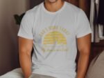 A person wearing a white t-shirt with yellow text reading "Can't Work Today My Arm is in a Cast" sits indoors.