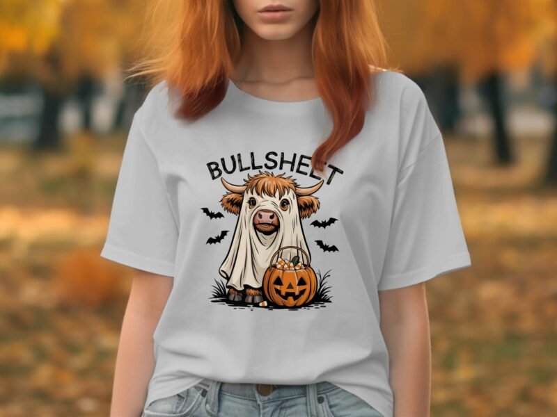A person wears a white t-shirt featuring a cartoon cow dressed as a ghost, with a jack-o'-lantern and bats.