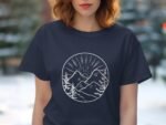 A woman wearing a navy t-shirt with a white mountain and sunset graphic, surrounded by blurred green foliage.