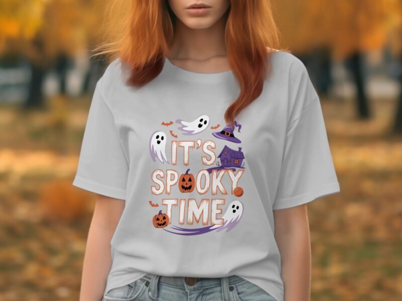 A woman with red hair wears a gray t-shirt featuring "It's Spooky Time" and Halloween graphics among fall trees.